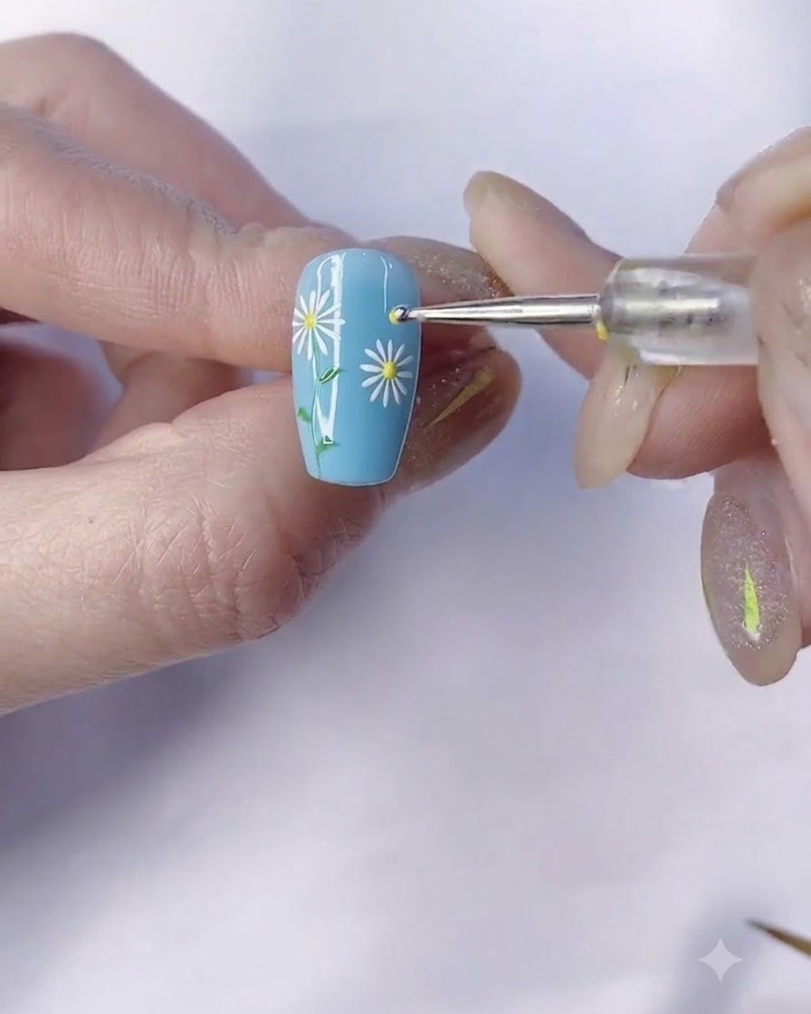 Un pincel fino dibujando un tallo verde hacia abajo desde la margarita en una uña con manicura azul.