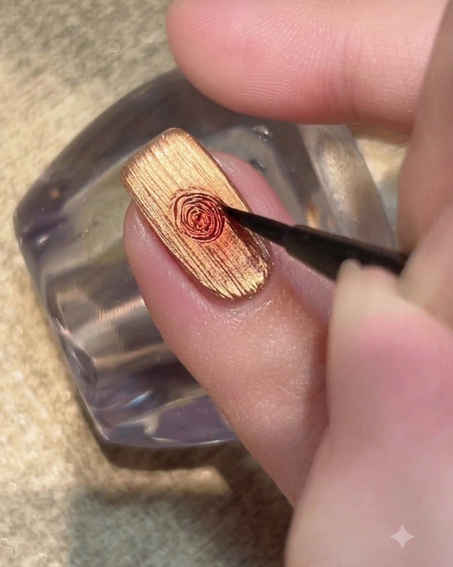 Un pincel fino de detalle difuminando una gota de gel rojo en un patrón de rosa circular sobre una uña dorada metálica.