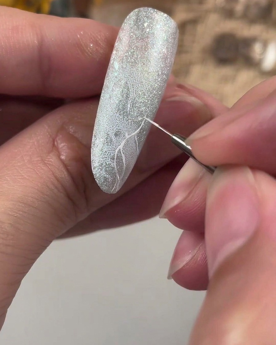 Un pincel de detalle fino dibujando una cruz guía blanca sobre una uña con purpurina texturizada.