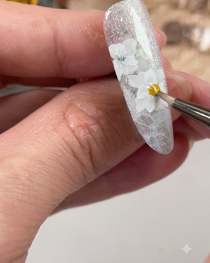 Un pincel de uñas aplicando una capa superior mate específicamente sobre los detalles de las flores blancas elaboradas en la uña.