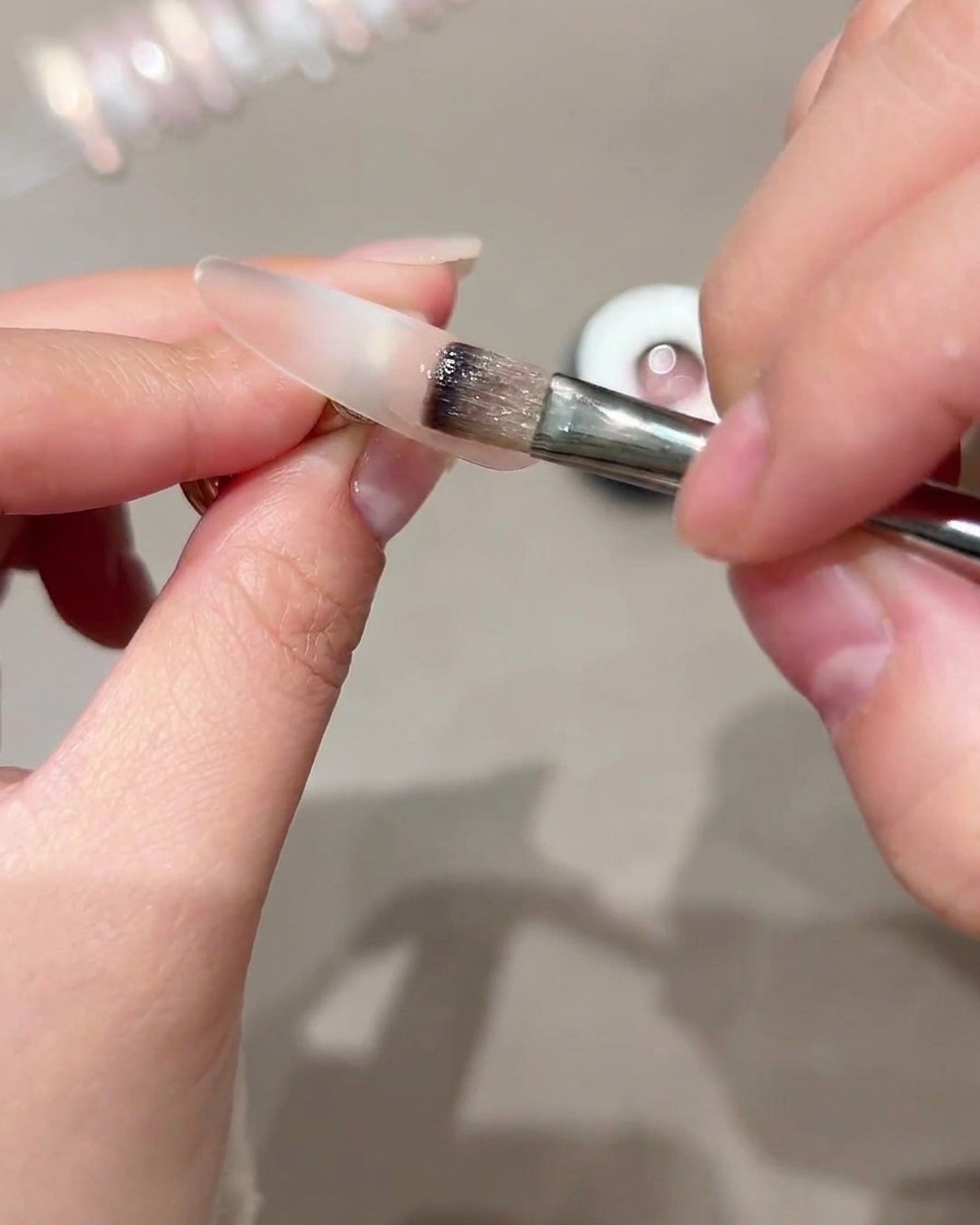 Un pincel aplicando gel ojo de gato en la porción exterior de una punta de uña transparente.