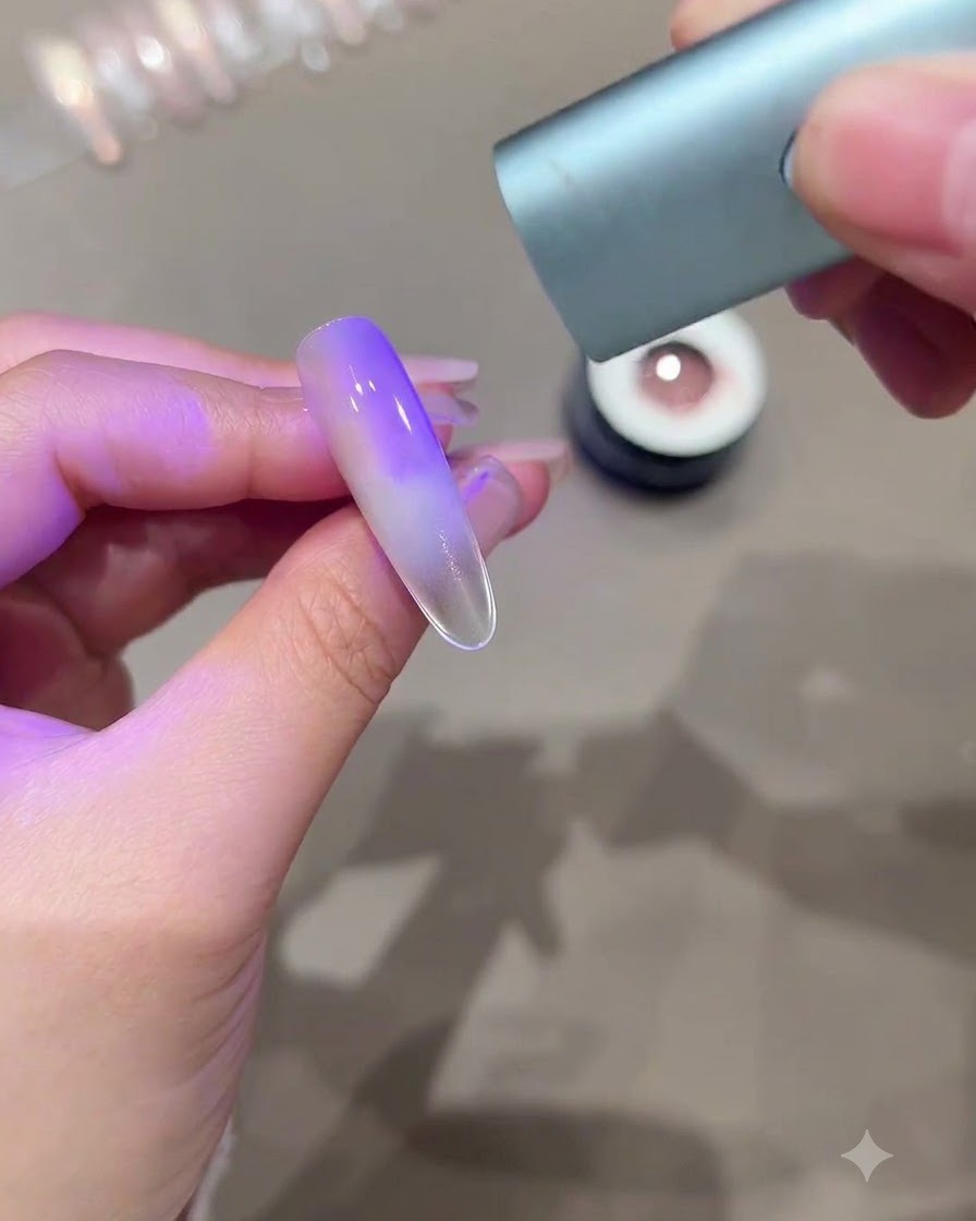 Una mano sosteniendo una mini luz de curado UV LED sobre una uña alargada que muestra un efecto de ojo de gato degradado en color púrpura.
