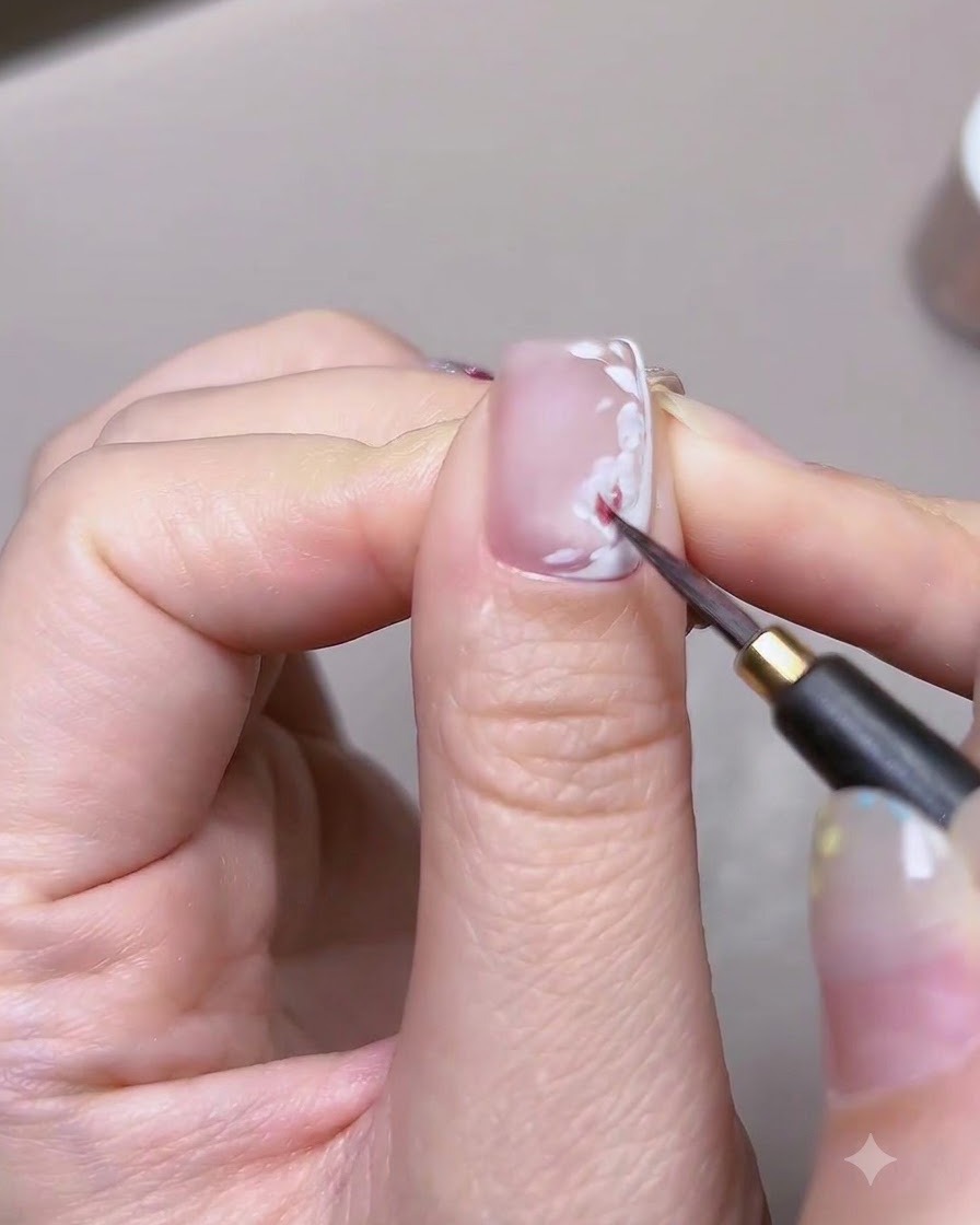 Un primer plano detallado de un pincel fino aplicando pintura de acuarela roja sobre la base de pétalos blancos de una uña con nail art de rosas.