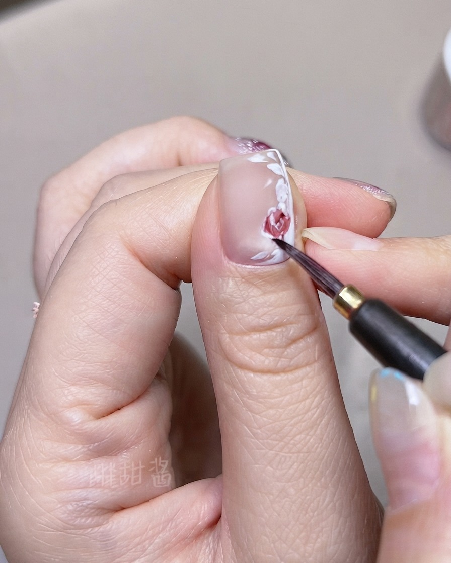 Un pincel fino superponiendo tonos de acuarela roja más oscuros en pétalos y hojas de rosa pintados individualmente en una uña.
