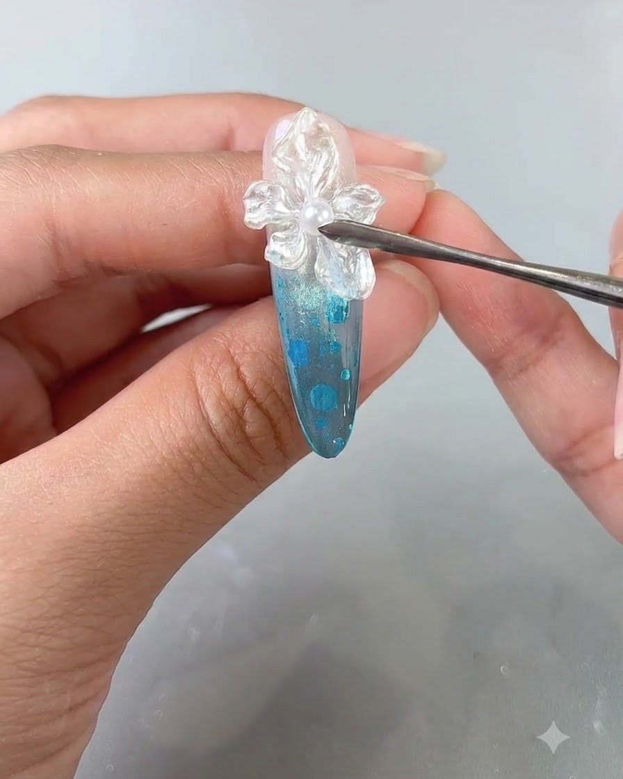 Un primer plano de un dedo manicurado recibiendo una aplicación final de capa superior transparente sobre un diseño de uñas con flores texturizadas.