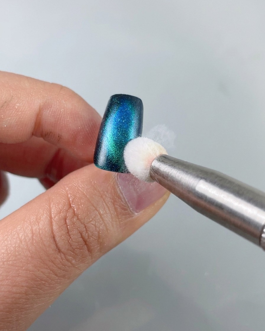 Una herramienta de esponja aplicando gel de estampado negro en el borde de una pieza de uña de ojo de gato verde azulado.