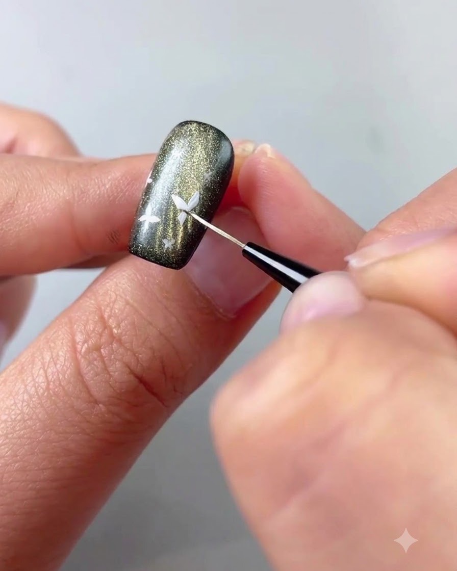 Una vista de cercanía de un pincel delineador fino aplicando gel blanco para pintar un detalle de mariposa pequeña en una uña oscura de ojo de gato.
