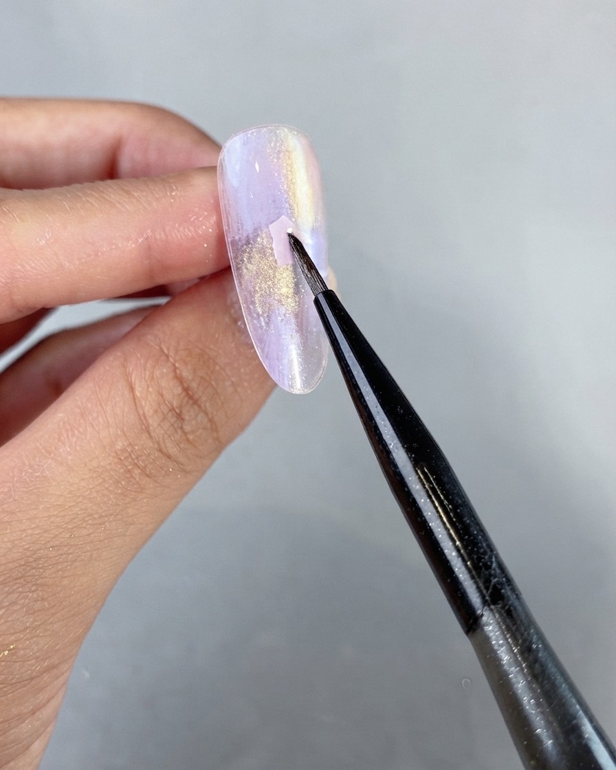 Un pincel delineador fino colocando delicadamente una pieza de concha rosa sobre la superficie de una uña de gel perlado morado sin curar.