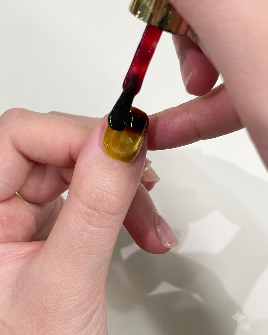 Un pincel de esmalte de uñas aplicando gel rojo en el borde superior de una uña con manicura de ojo de gato amarillo.
