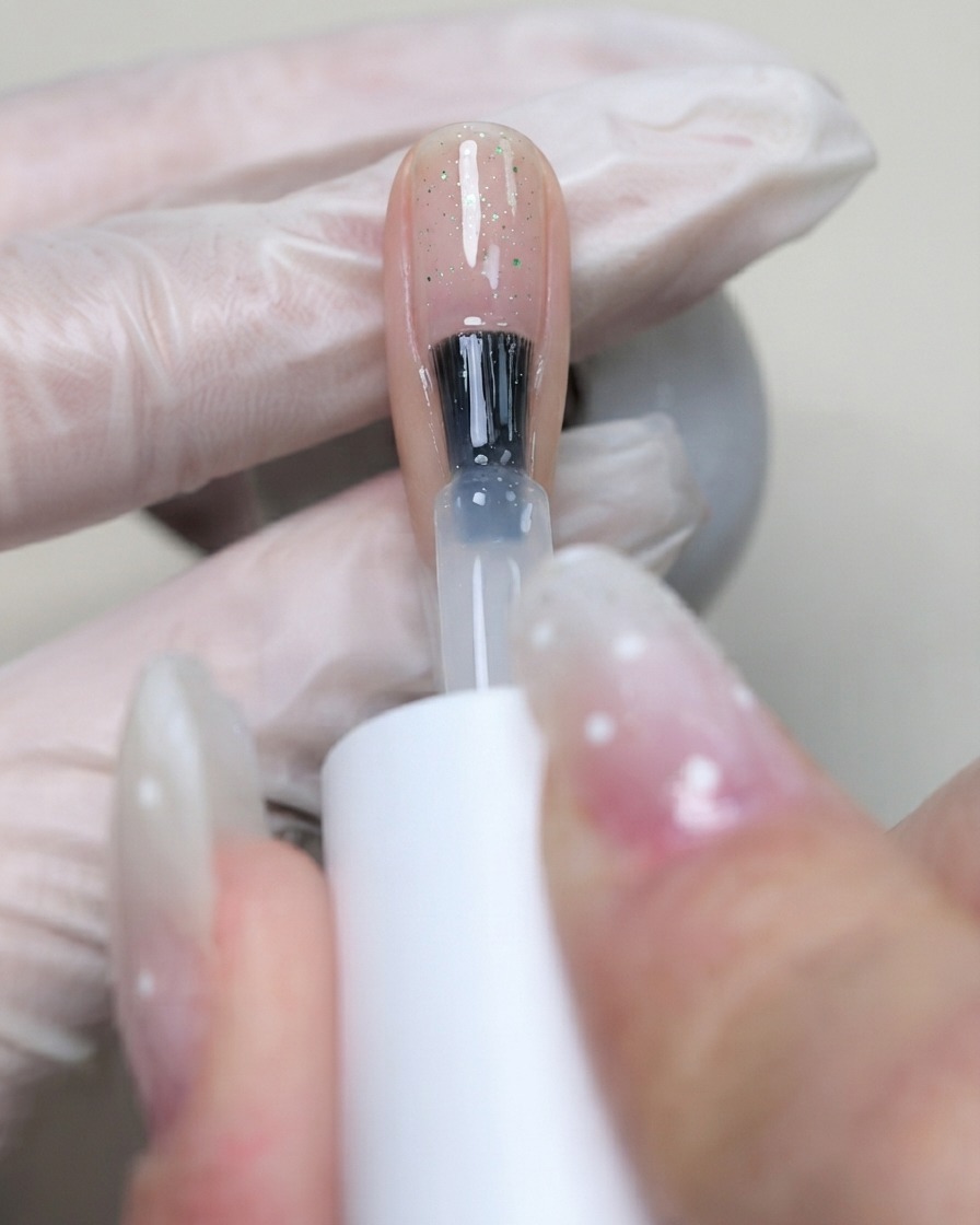 Un pincel aplicando gel base flexible C7 transparente sobre la superficie de la uña para fijar las lentejuelas colocadas.
