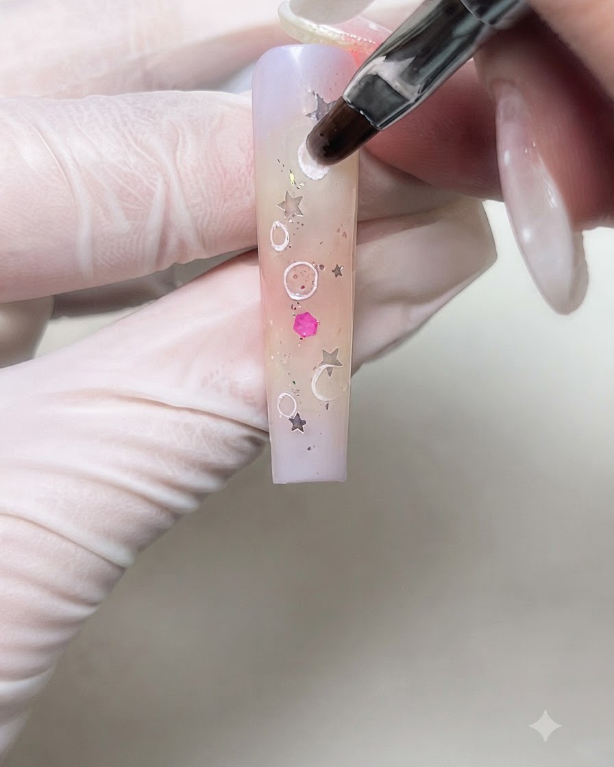 Un pincel de nail art difuminando suavemente los bordes internos de los círculos de crema blanca en una uña con degradado.
