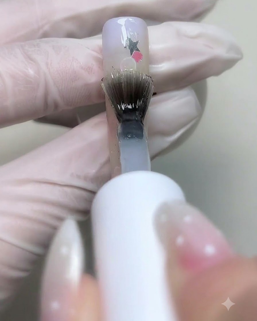 Un pincel aplicando gel base transparente sobre uñas decoradas con lentejuelas de estrellas y burbujas.