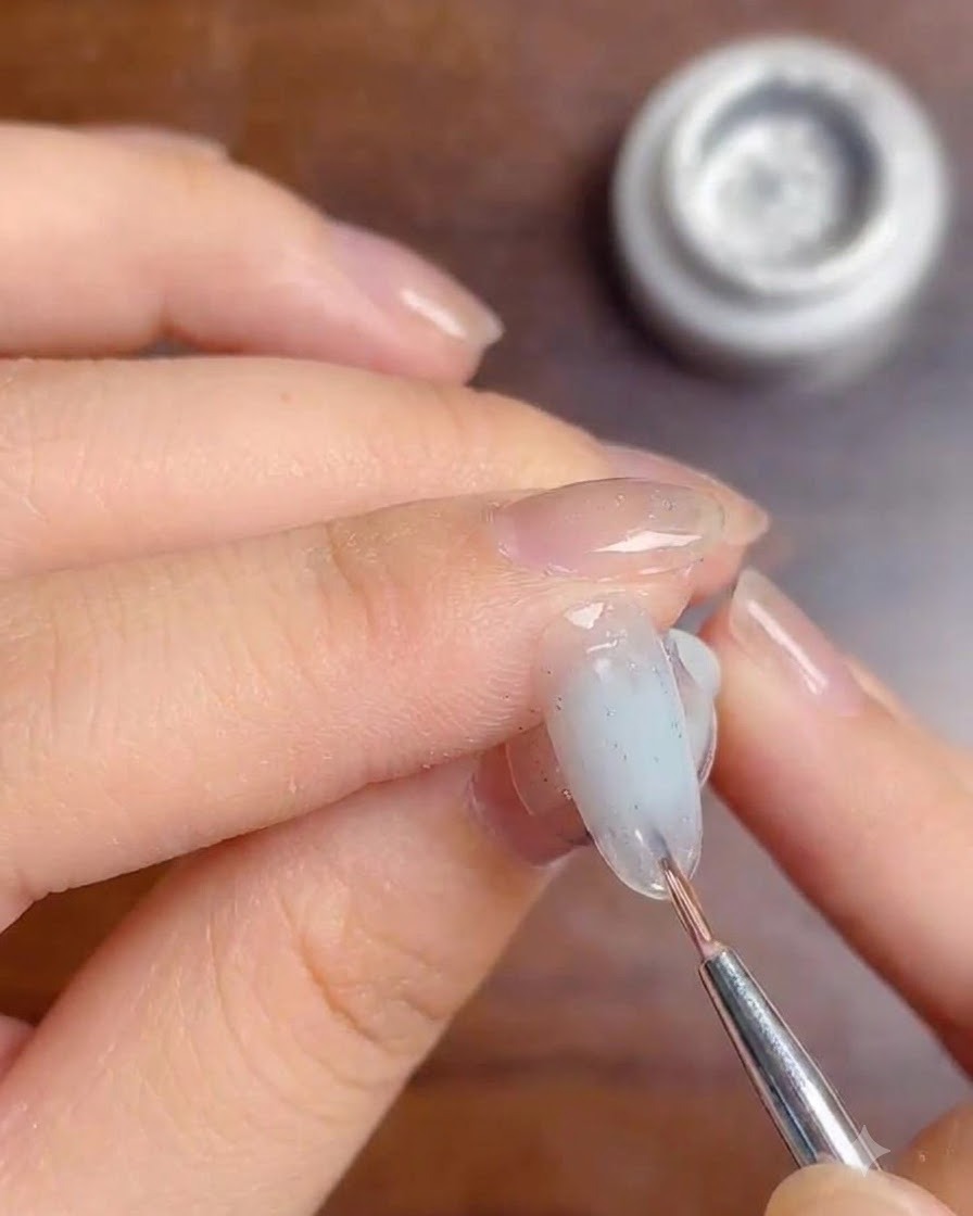 Un pincel fino aplicando una capa de gel de acabado transparente sobre las líneas de purpurina plateada en una uña con degradado blanco.