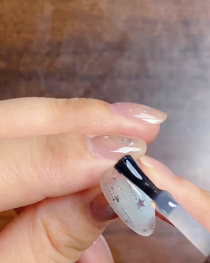 Un pincel de uñas aplicando una capa superior brillante final sobre una uña con degradado lechoso decorada con purpurina plateada y pegatinas de estrellas.