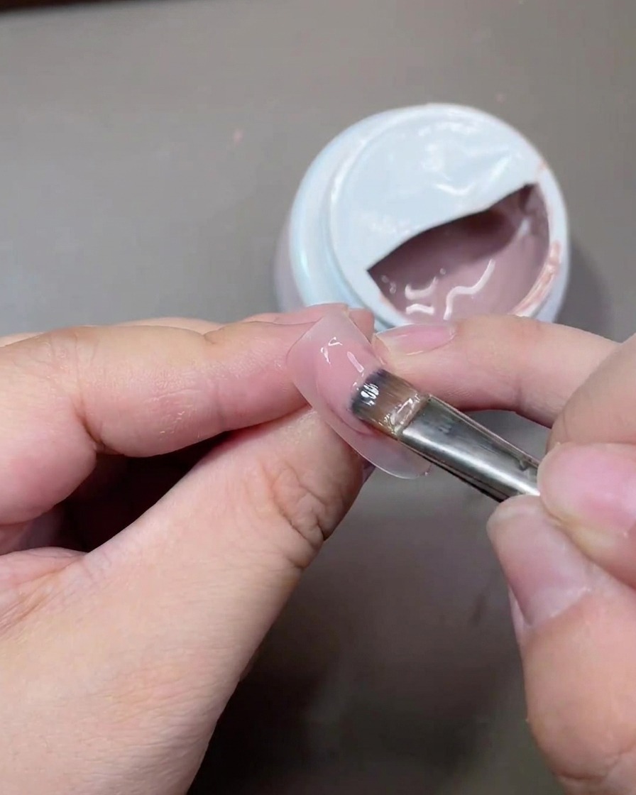Un pincel de gel plano aplicando una capa suave de esmalte nude translúcido sobre la superficie de una uña.
