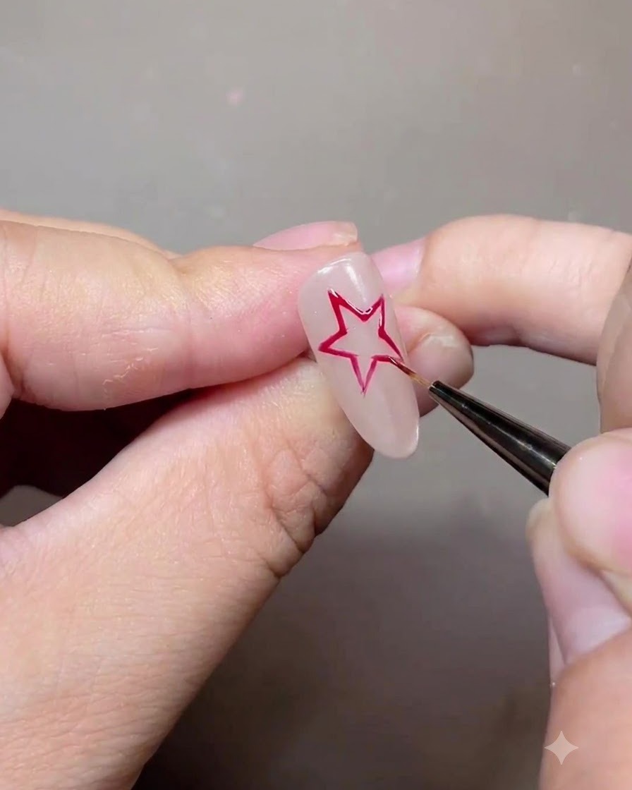 Un pincel delineador fino dibujando el contorno de una estrella roja de cinco puntas en una uña de gel nude.