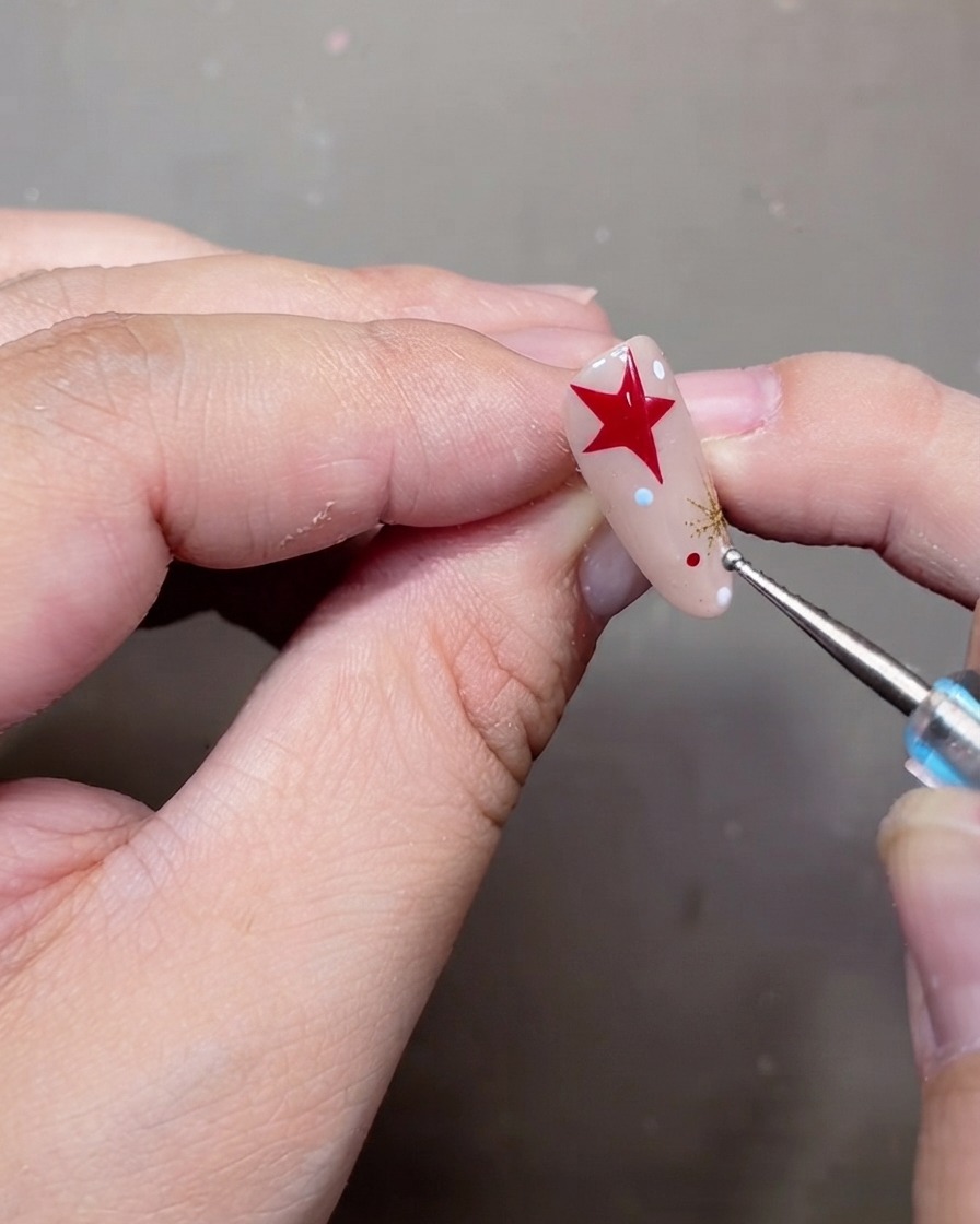 Una herramienta punteadora colocando pequeños lunares blancos y rojos en una uña de gel nude decorada.