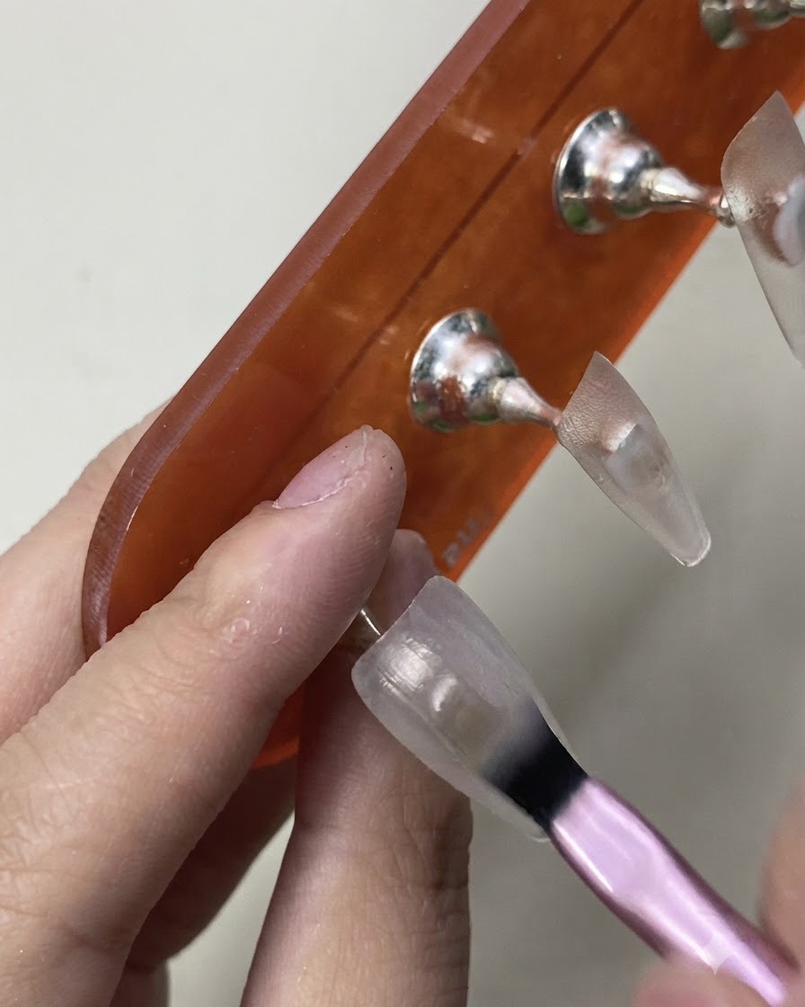 Un pincel aplicando cuidadosamente la capa inicial de esmalte en gel blanco lechoso translúcido sobre un tip de práctica.