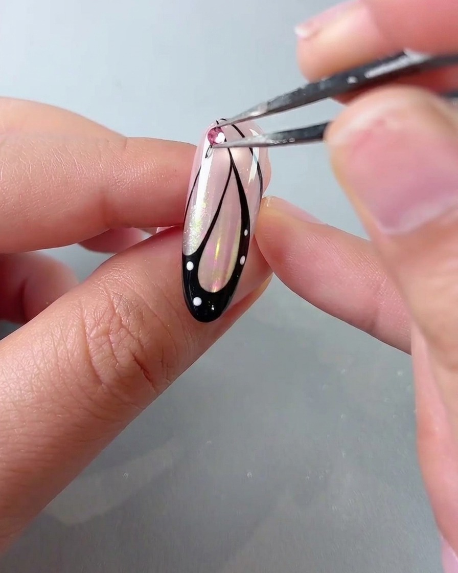 Un par de pinzas colocando un estrás de cristal rosa en la parte superior de un diseño de uña con alas de mariposa.