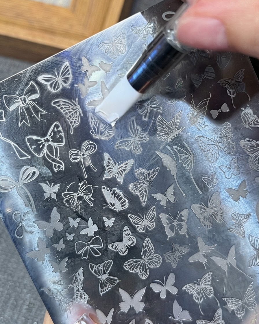 Una herramienta esparciendo gel de estampado blanco sobre un patrón de mariposa en una placa de estampación metálica.