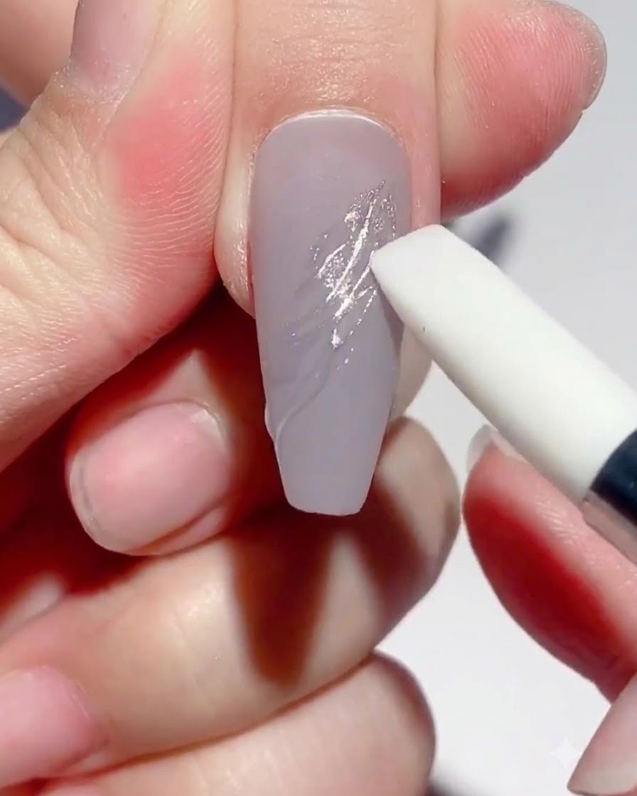 Una herramienta con punta de silicona frotando polvo espejo plateado sobre líneas de gel curadas en una uña tono topo.