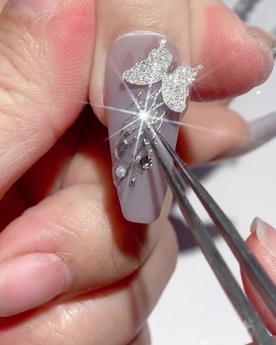 Unas pinzas sosteniendo un dije mientras se aplica pegamento para pedrería alrededor de las decoraciones de uñas.