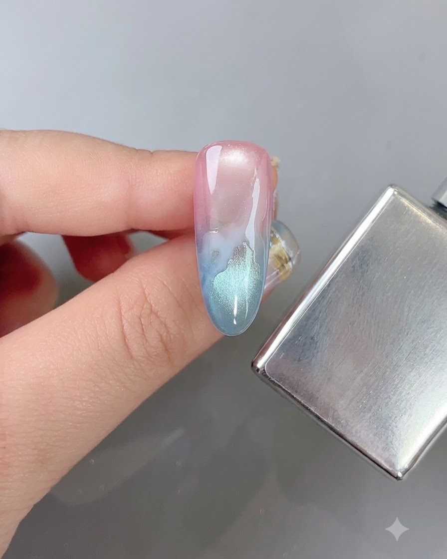 Un pincel de uñas difuminando suavemente los bordes de un patrón de nube blanca en una uña degradada multicolor junto a un bastón magnético.