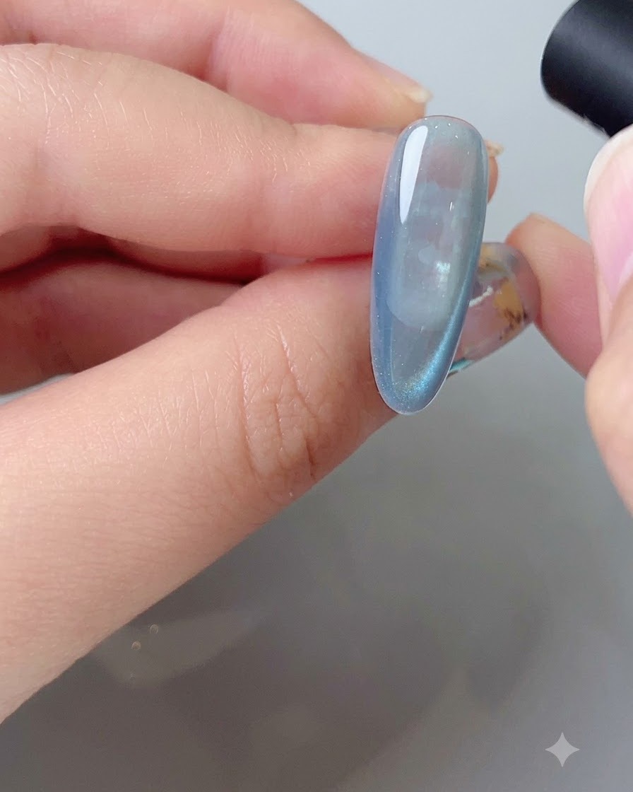 Una herramienta magnética moldeando el arco de reflexión lateral en una uña de gel azul con efecto de ojo de gato.