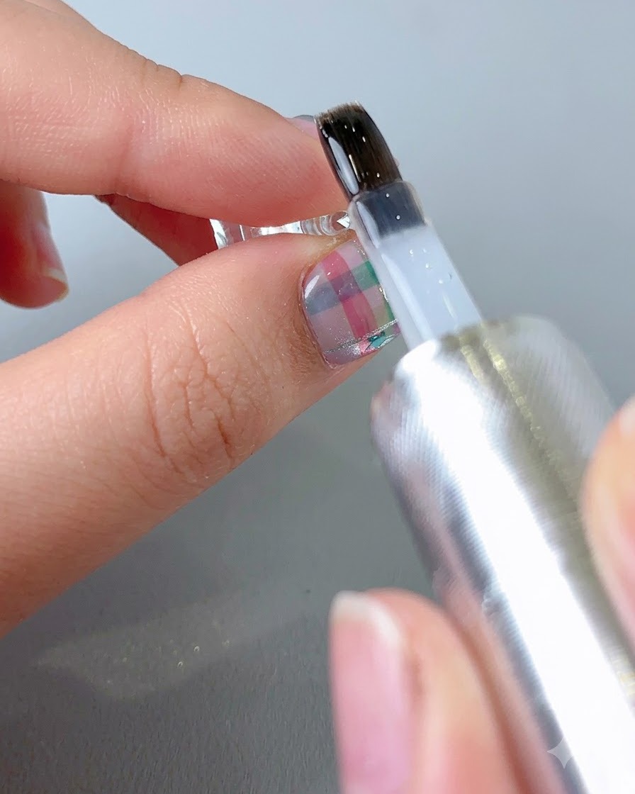 Un pincel aplicando un top coat transparente y brillante en la superficie de una uña con patrón terminado.