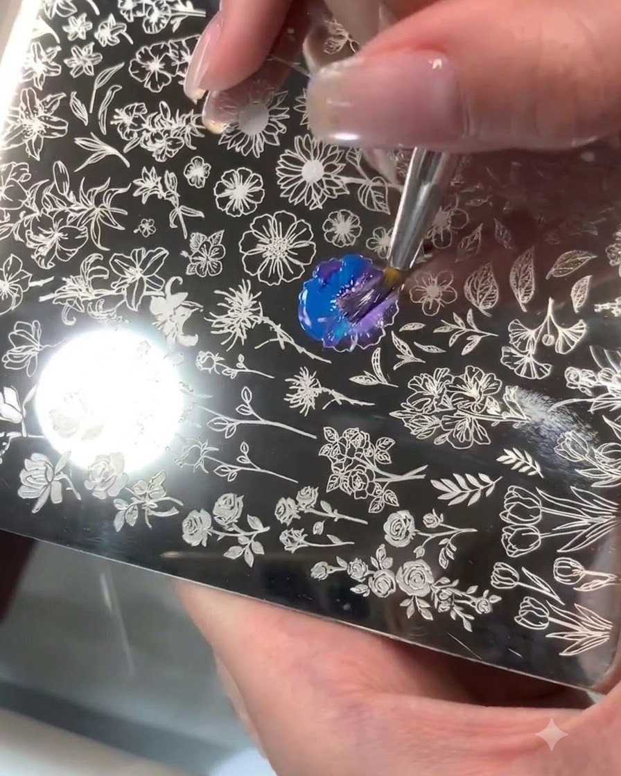 Una placa de acero para estampado con un patrón floral grabado donde se mezclan gel azul y púrpura con un pincel.