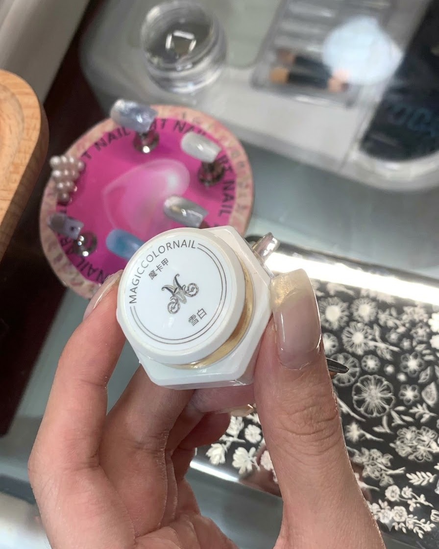 Una mano sosteniendo un producto de estampado blanco en botella con caracteres asiáticos y una placa de estampado de uñas de fondo.