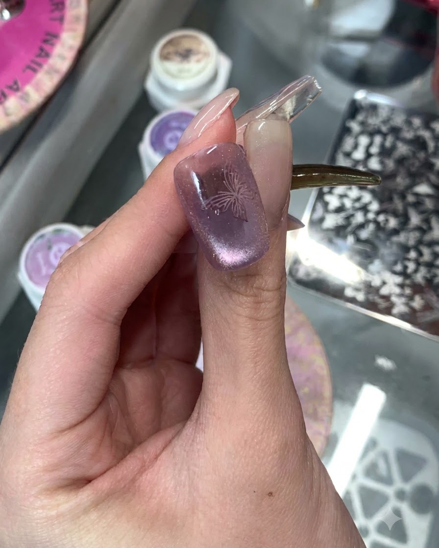 Un primer plano de una uña manicurada con efecto ojo de gato púrpura con un delicado diseño de mariposa estampado encima.