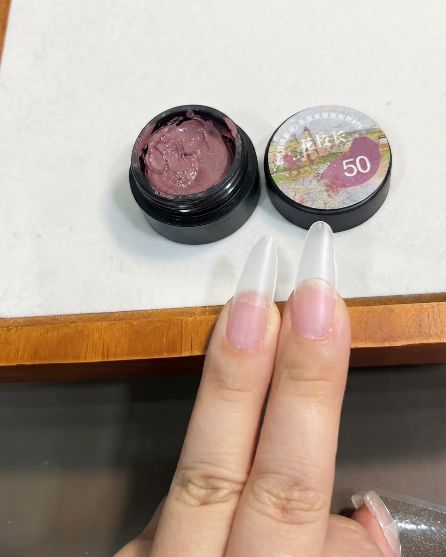 Un pincel sosteniendo una pequeña cantidad de gel sólido rosa pasta de frijol cerca de la punta de una extensión de uña transparente.