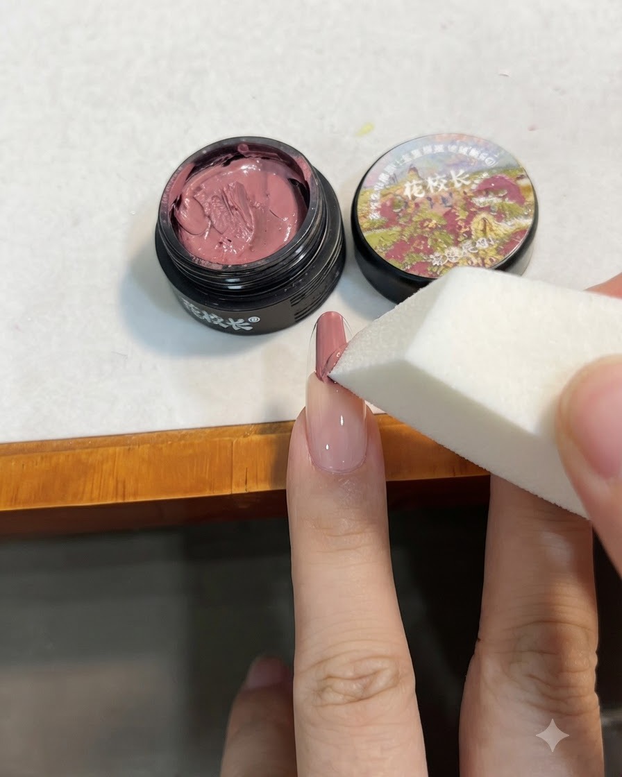 Una esponja de maquillaje blanca dando golpecitos sobre el gel rosa en la uña para difuminarlo hacia abajo en un degradado.
