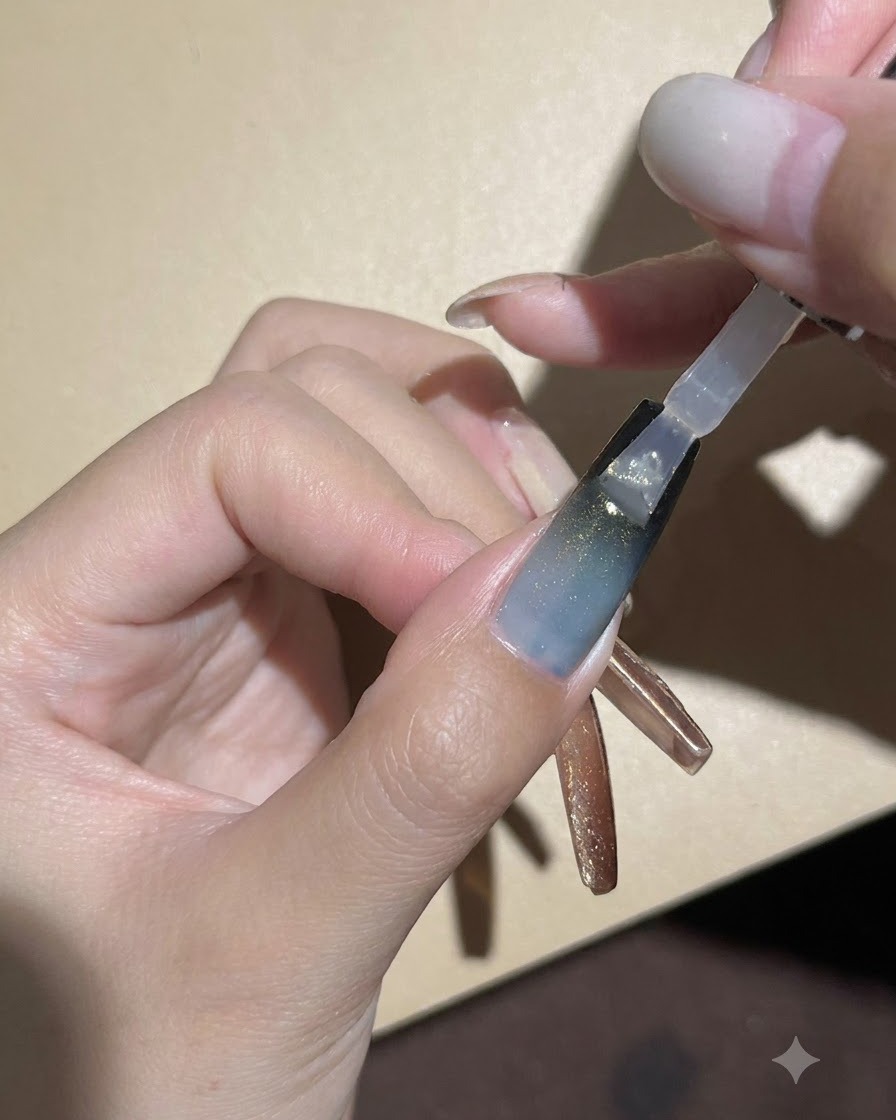 Un pincel de uñas aplicando una capa superior brillante y transparente sobre la uña de gel con degradado oscuro y ojo de gato.