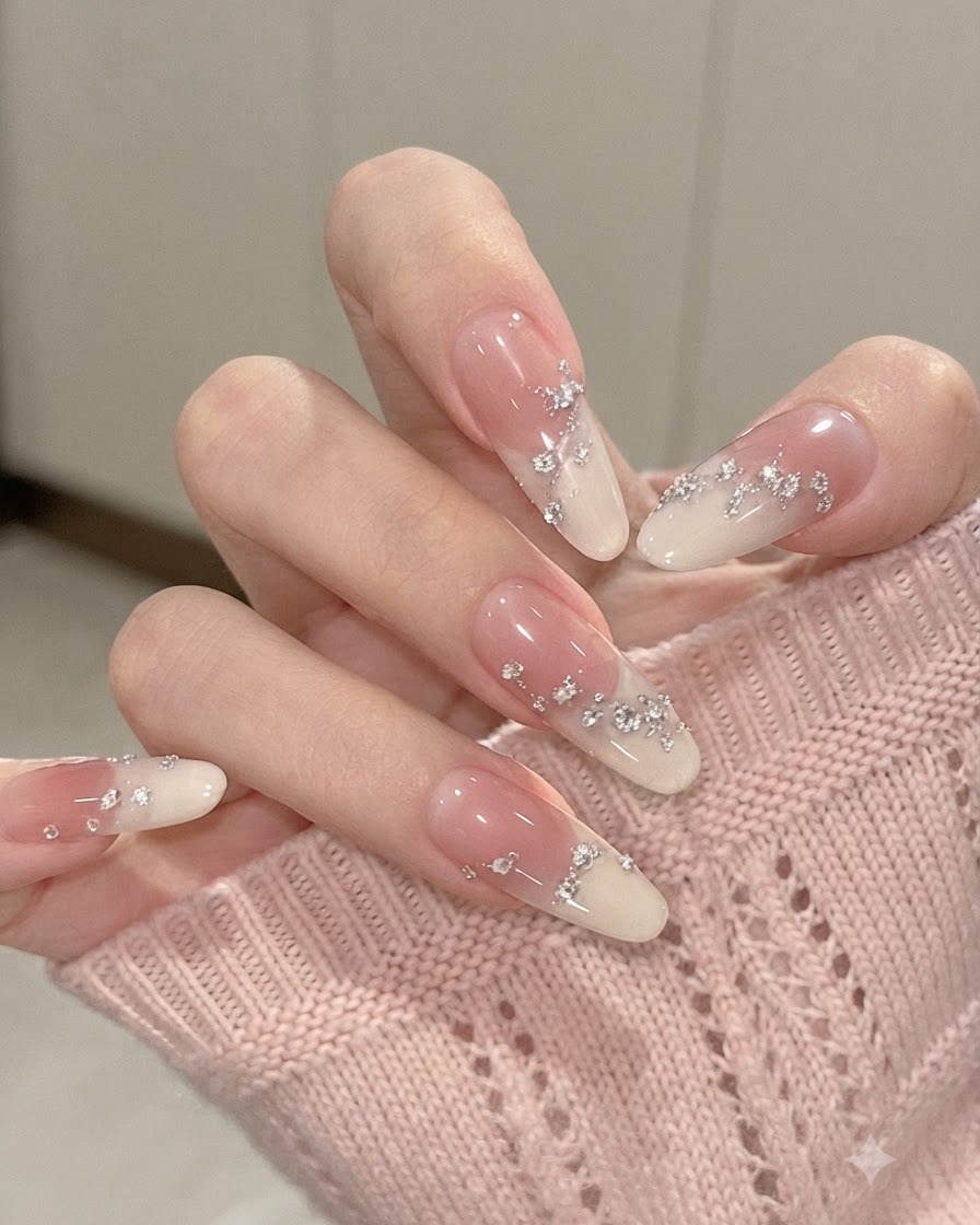 Vista de cerca de uñas degradadas en forma de almendra con los dedos flexionados, resaltando las láminas plateadas y los pequeños diamantes de imitación.