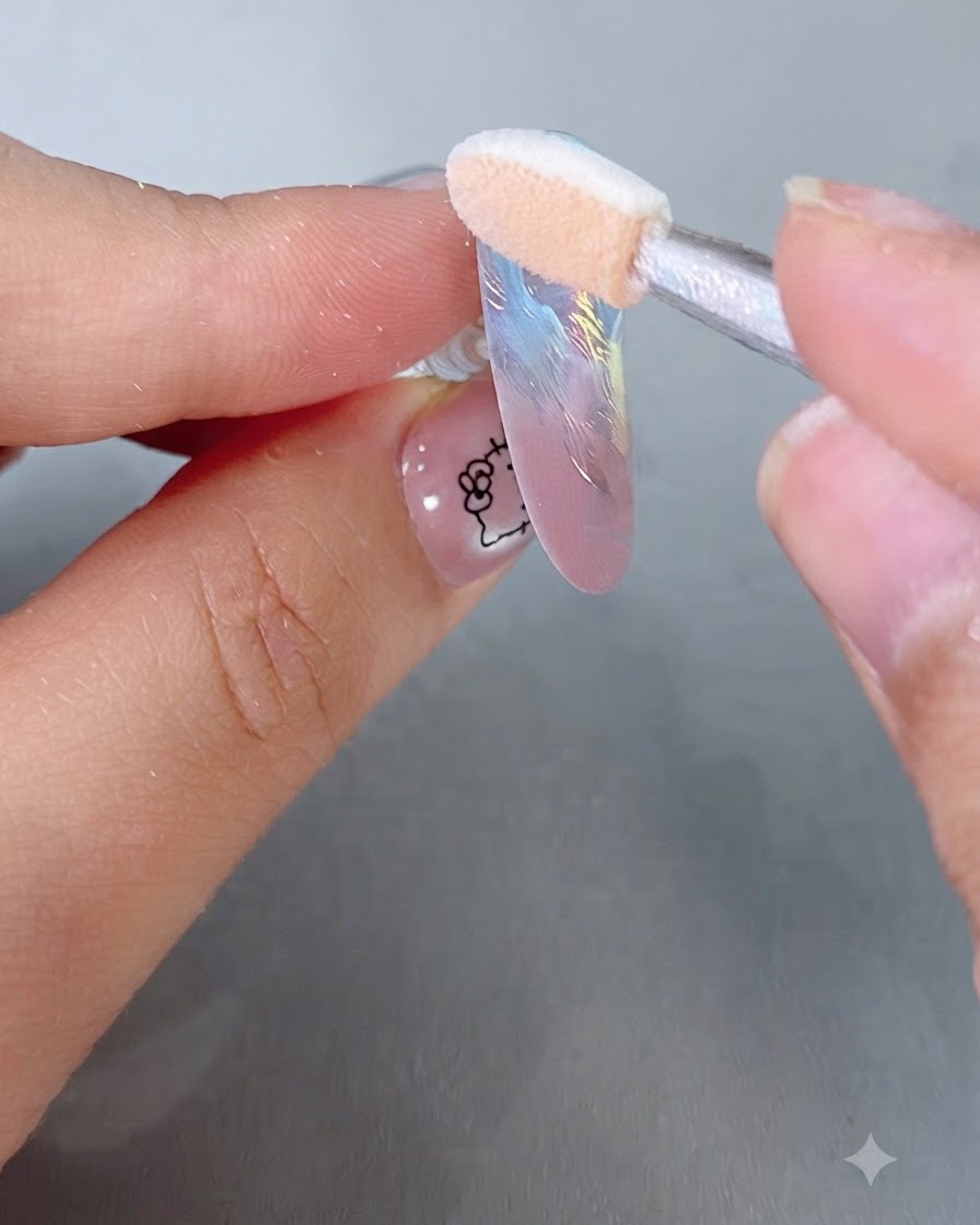 Un aplicador de esponja frotando polvo espejo mágico iridiscente sobre una uña postiza en tonos pastel.