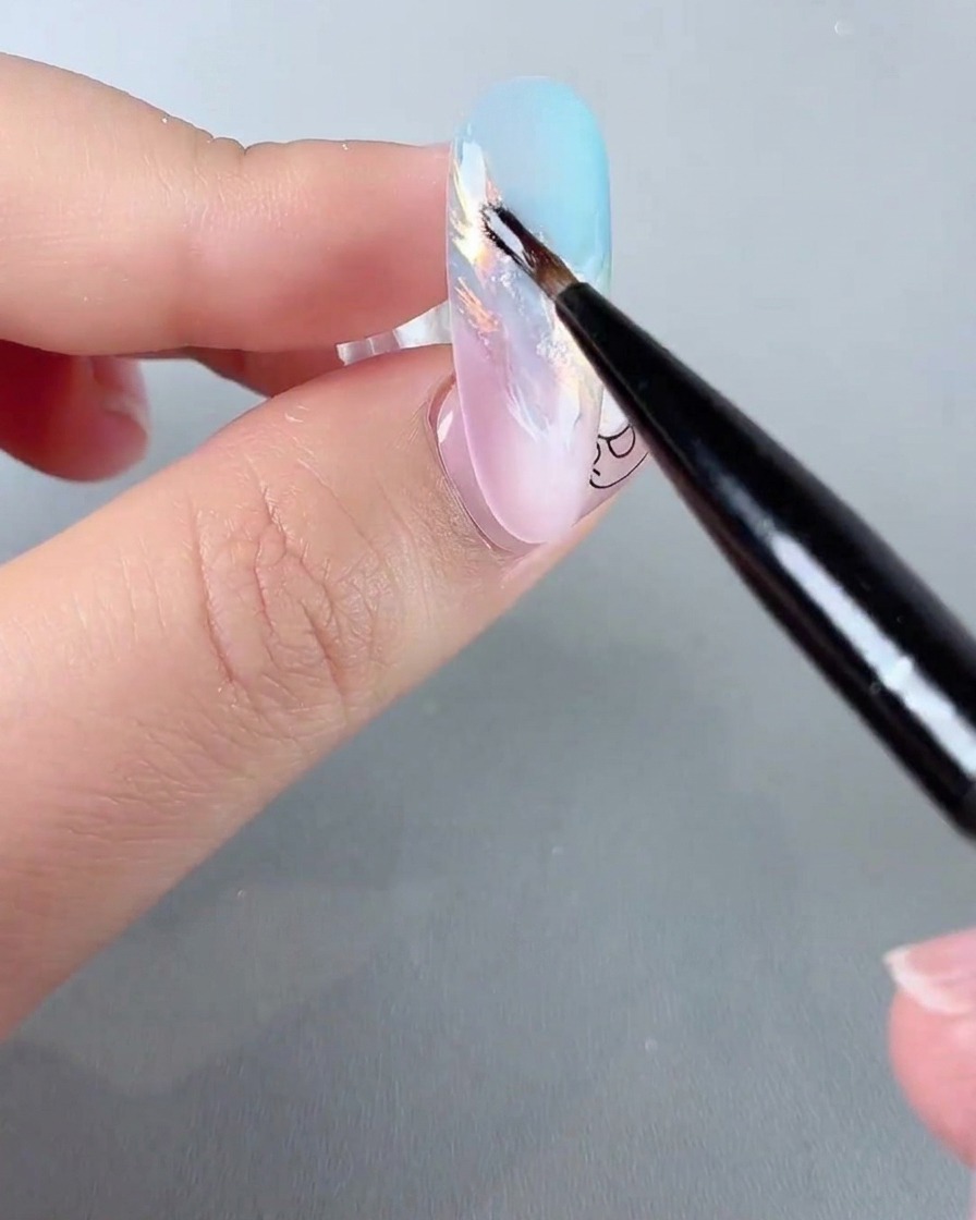 Un pincel fino aplicando detalles de glitter iridiscente brillante sobre una uña con degradado pastel.