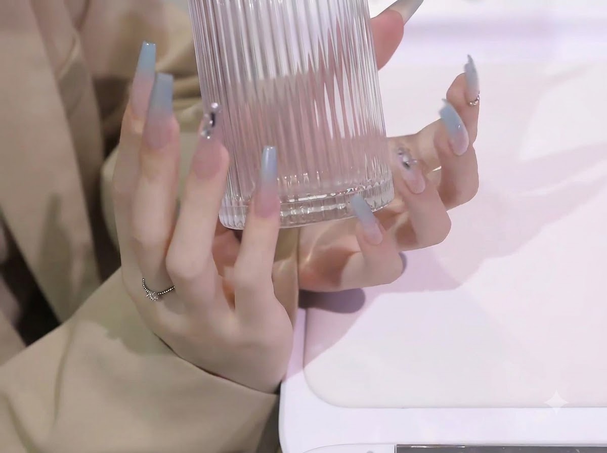 Ambas manos levantando un recipiente de vidrio transparente acanalado, mostrando la longitud y la curvatura de las extensiones de uñas con degradado azul.