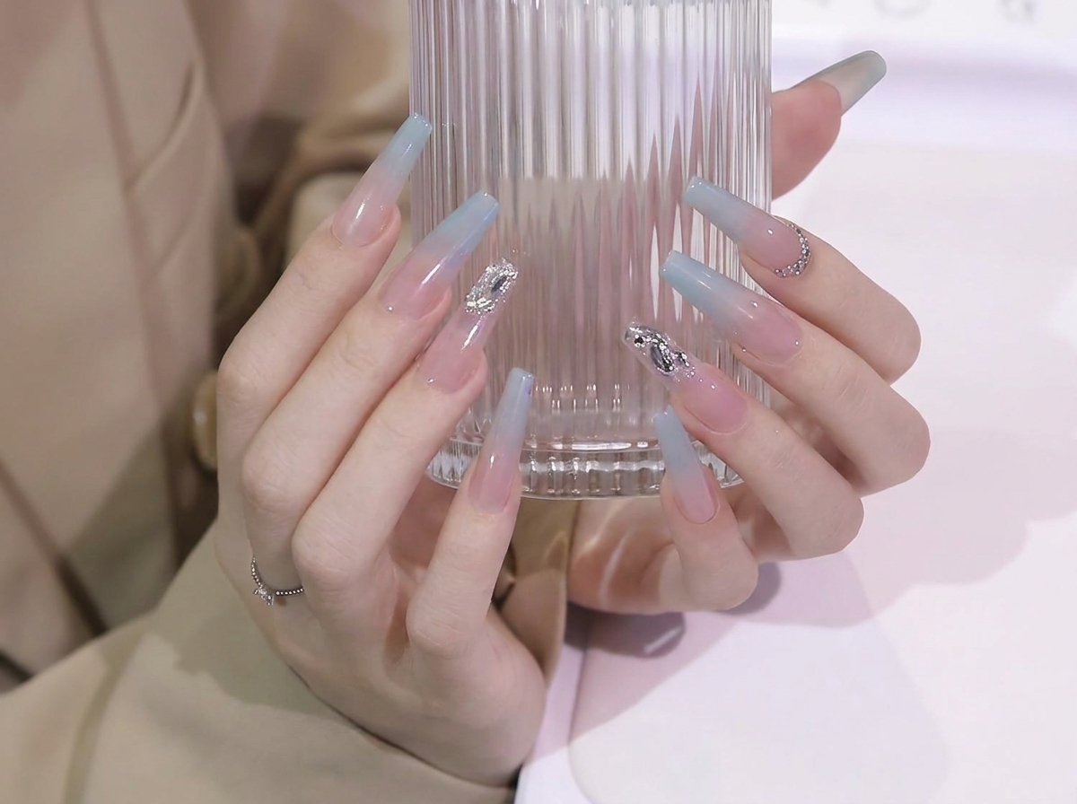 Manos ajustando el agarre en un vaso de vidrio para mostrar la suave transición del degradado de azul a nude en uñas coffin largas.