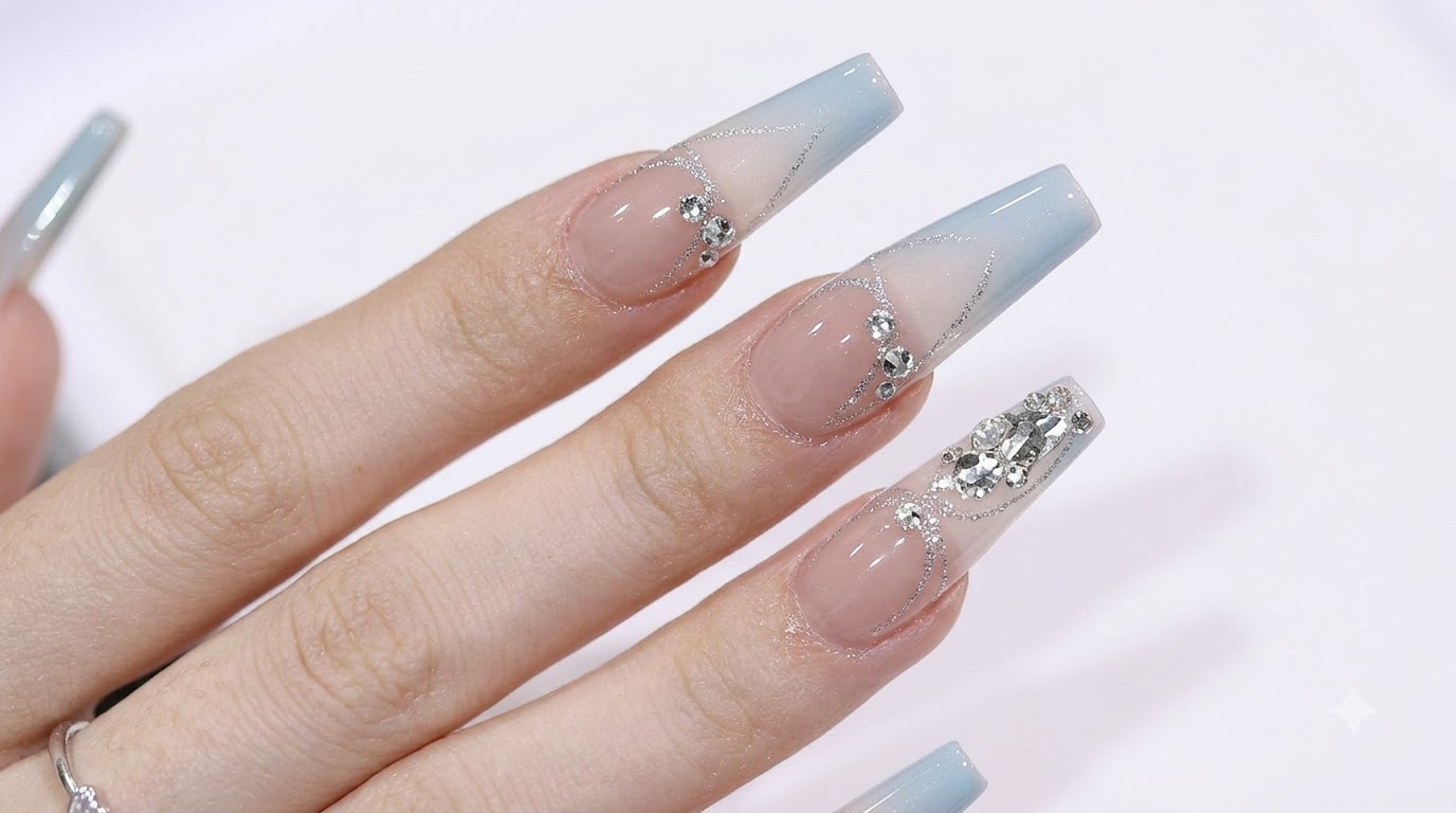 Uñas largas en forma coffin con un suave degradado de azul a nude, decoradas con pedrería brillante y detalles de purpurina plateada.