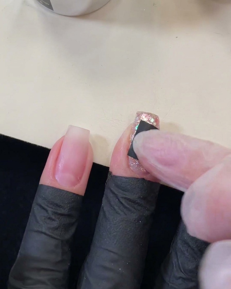 Un dedo presionando una pequeña pegatina cuadrada negra con un recorte en forma de estrella sobre una uña brillante con lentejuelas.