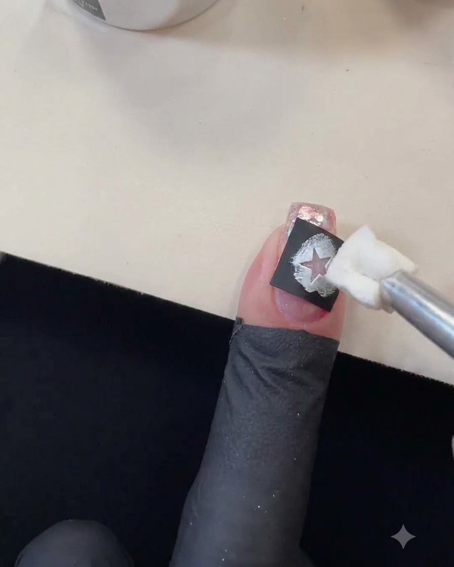 Un aplicador de esponja aplicando gel de color blanco sobre el recorte de una plantilla de estrella negra en una uña con purpurina.