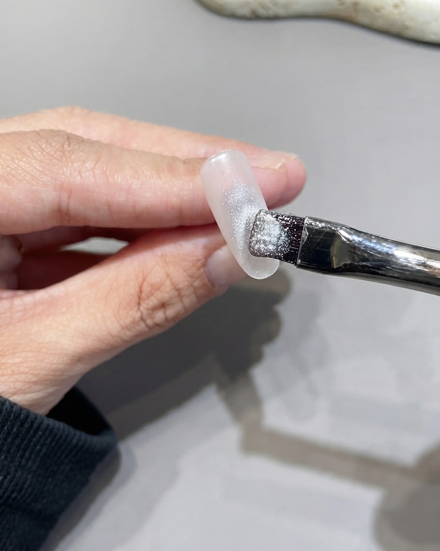 Un pincel plano aplicando gel ojo de gato plateado brillante sobre la superficie de una uña.