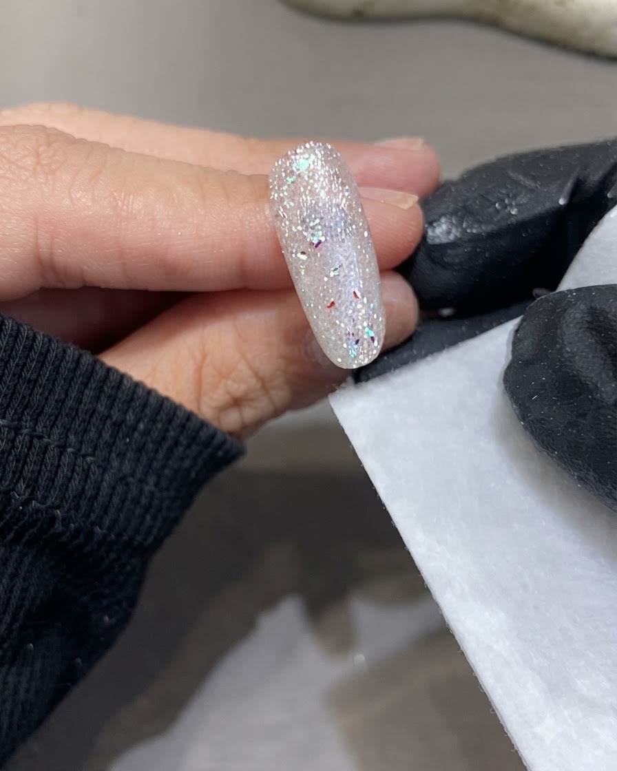 Una mano con guantes sosteniendo una toallita sin pelusa junto a la superficie curada de una uña brillante.