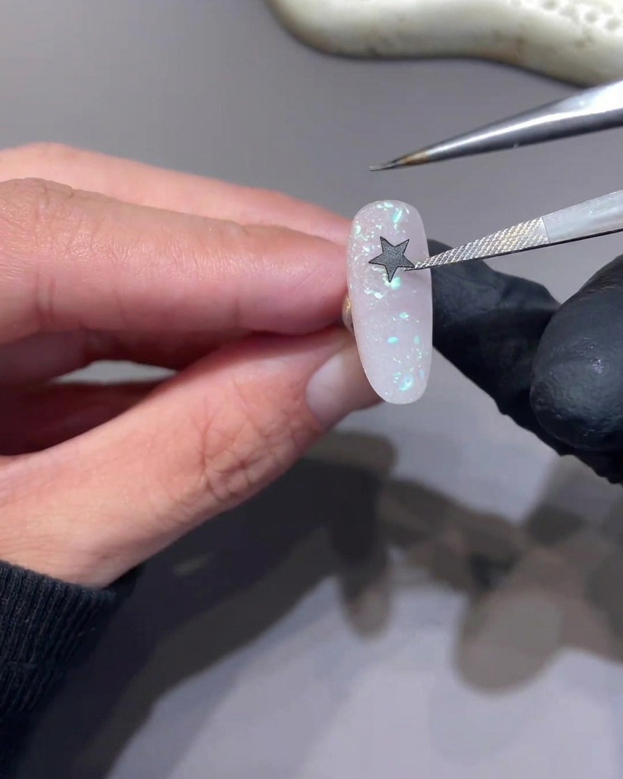 Unas pinzas colocando una pegatina negra en forma de estrella sobre la superficie de una uña rosa con purpurina.