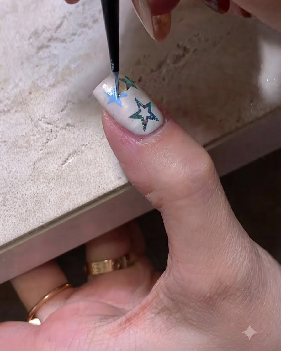 Primer plano de un pincel fino de nail art dibujando el contorno azul de una estrella en una uña arreglada.