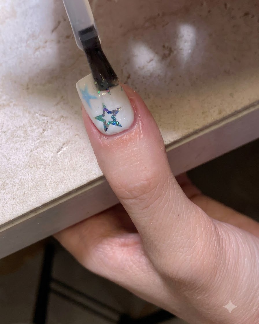 Un pincel aplicando un top coat transparente y brillante sobre una uña decorada con estrellas y purpurina.