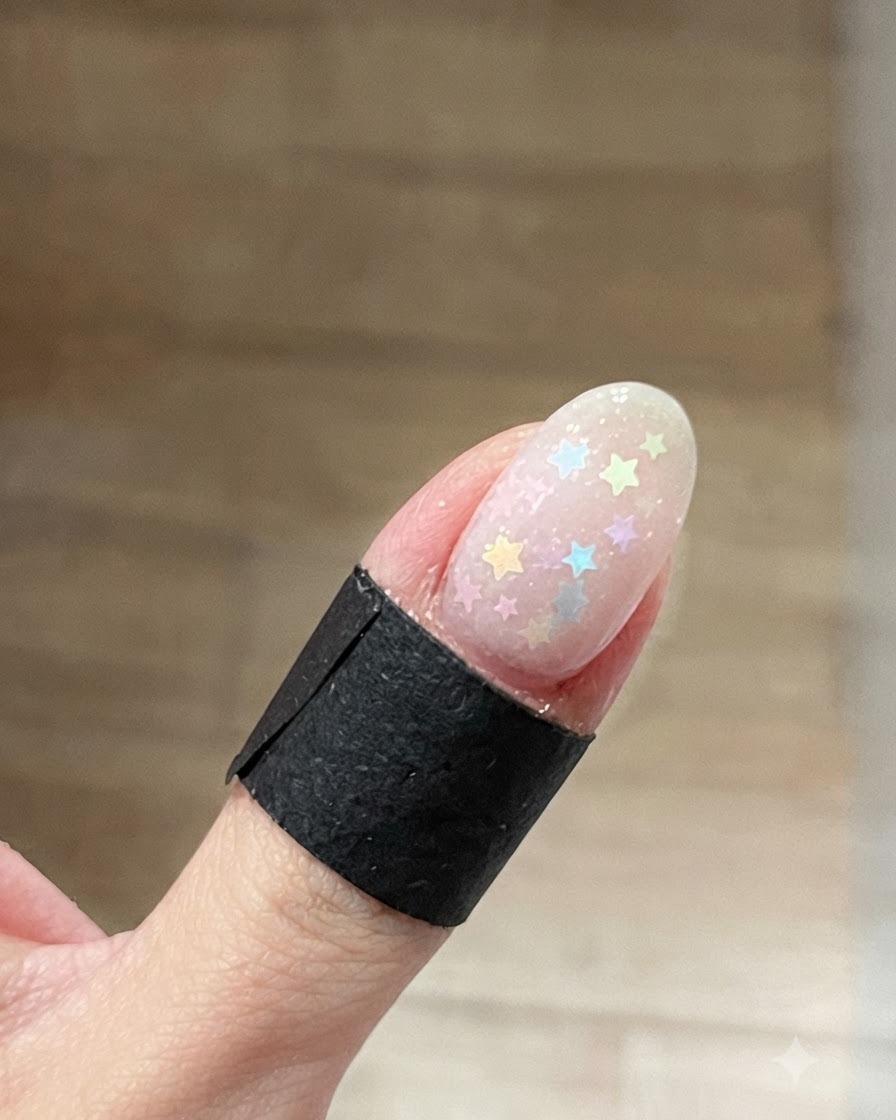Primer plano de una uña ovalada con glitters de estrellas multicolores esparcidos sobre un gel base húmedo y transparente.