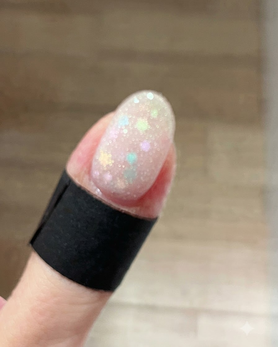 Primer plano de una uña cubierta con un gel brillante blanco escarchado sobre glitters de estrellas en tonos pastel.